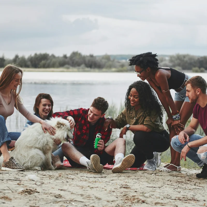 A group of friends sits on the beach, smiling and gathered around a fluffy white dog.